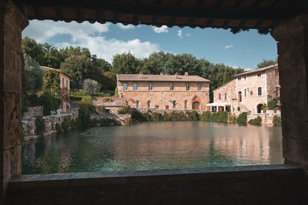 Vue sur la place d’eau thermale de Bagno Vignoni, entourée de bâtiments en pierre typiques de la Toscane, capturée depuis une loggia ombragée